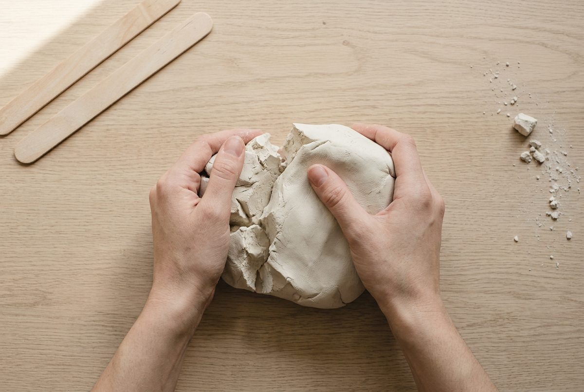 Hands kneading a lump of off-white air dry clay on a pale oak table